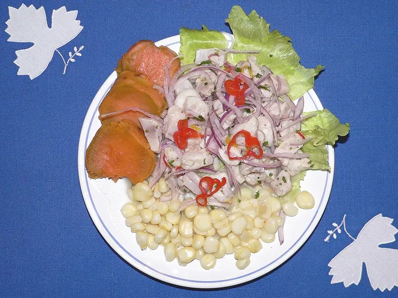 Ceviche of white fish with red onion and red chili rings on lettuce, served with sweet potato slices and large corn kernels on a blue cloth