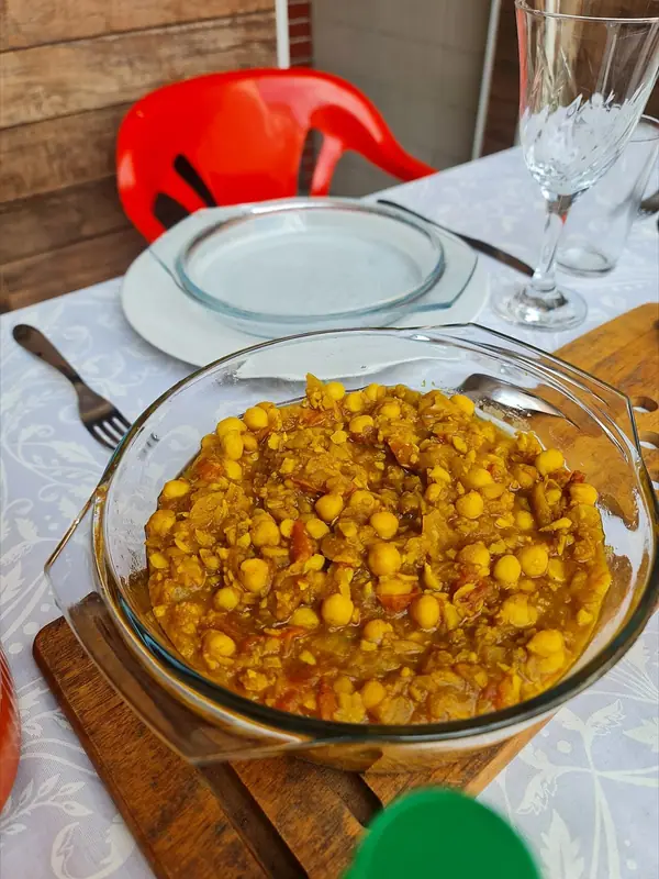 Glasform mit goldgelbem Kichererbsen-Curry mit Tomatenstückchen auf einem Holzbrett, auf einer weißen gemusterten Tischdecke neben einem leeren Teller