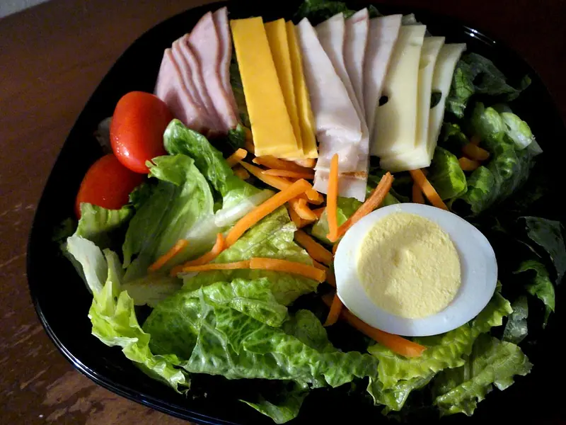 Romainesalat i en sort skål med rækker af skinkeskiver, kalkun, cheddar og schweizerost, revet gulerod, cherrytomater og et halveret æg