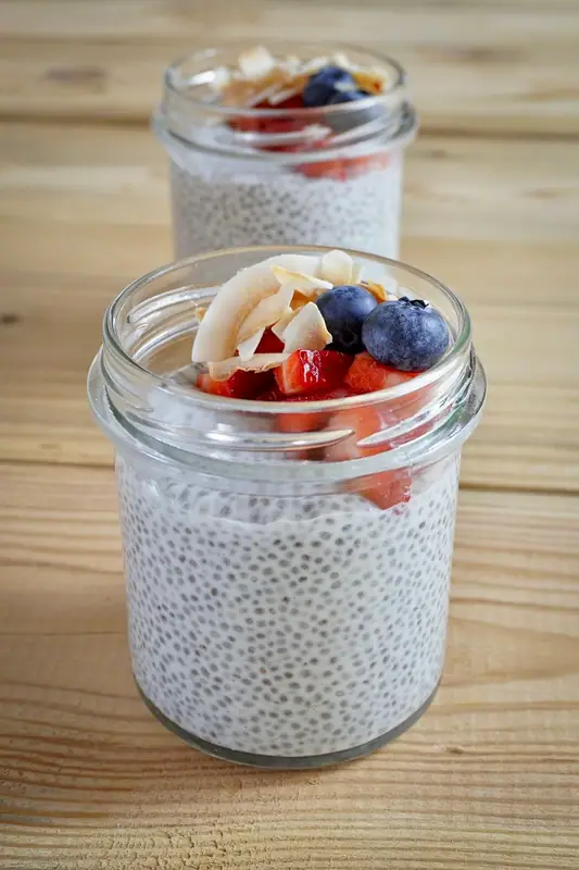 Two glass jars of chia pudding on a wooden table, topped with diced strawberries, blueberries and toasted coconut flakes.