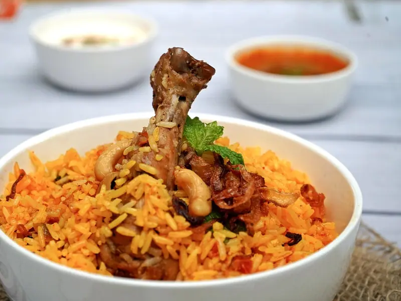 Oranžově zbarvená rýže biryani v bílé misce s kuřecí paličkou, kešu oříšky, smaženou cibulkou a mátou, v pozadí raita a kari.