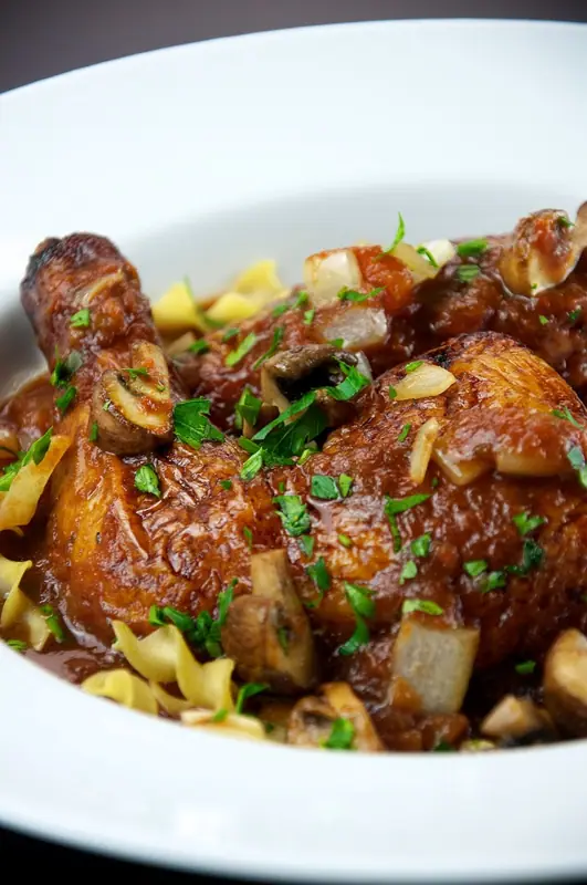 Browned bone-in chicken legs in a rich tomato sauce with mushrooms and onion, sprinkled with parsley over egg noodles in a white bowl.