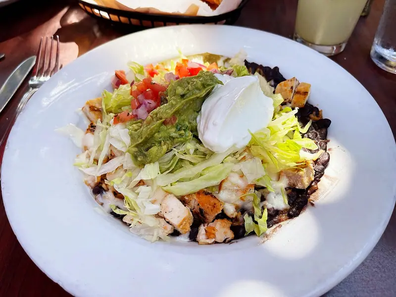 Bowl som på restaurant med grillet kylling, sorte bønner og smeltet ost, toppet med strimlet salat, guacamole, creme fraiche og pico de gallo