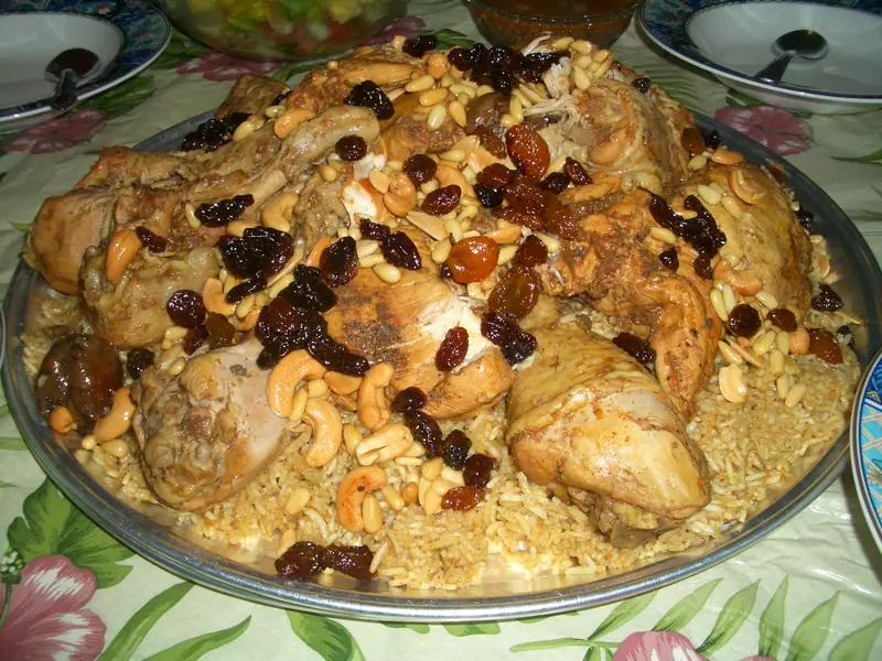 Große Metallplatte mit gewürztem Reis, belegt mit Hähnchenteilen mit Knochen, Rosinen, Cashewkernen und Pinienkernen, auf einer geblümten Tischdecke.