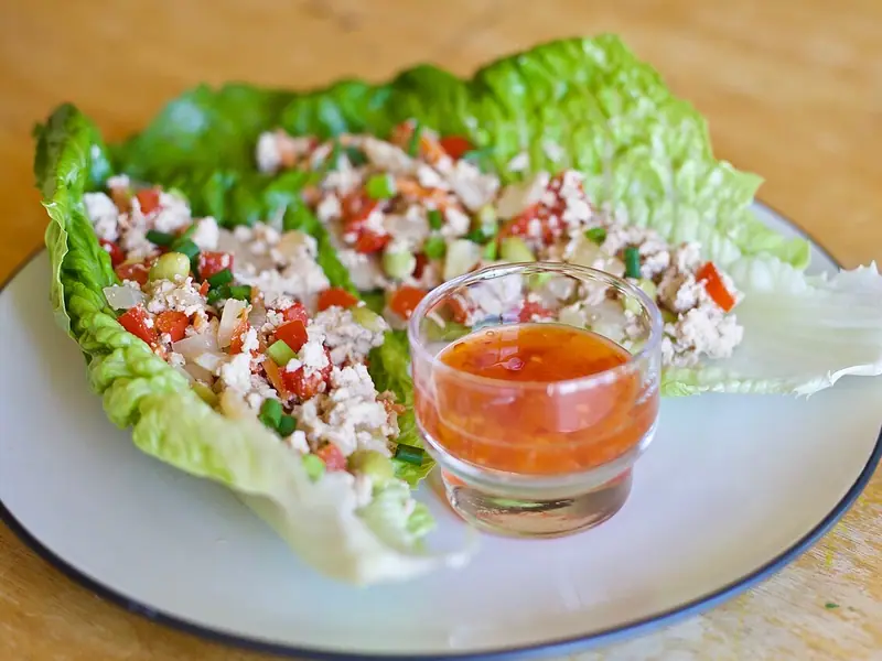Romainesalatblade fyldt med hakket kylling, tomat i tern, løg og forårsløg, med et lille glas sød chilisauce på en tallerken.