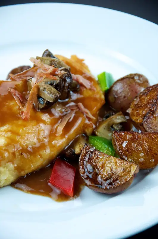 Filé de frango dourado em molho escuro coberto com cogumelos e presunto cru crocante, acompanhado de batatas vermelhas e pimentões assados em um prato branco.