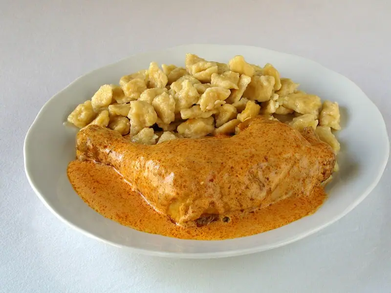 A bone-in chicken leg quarter in creamy orange paprika sauce beside a pile of small egg dumplings on a white plate.