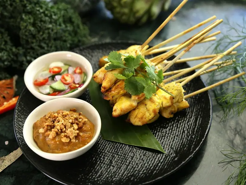 Gule grillede kyllingsatayspyd på et bananblad med koriander, en skål peanøttsaus toppet med nøtter og en agurk- og chilisalat