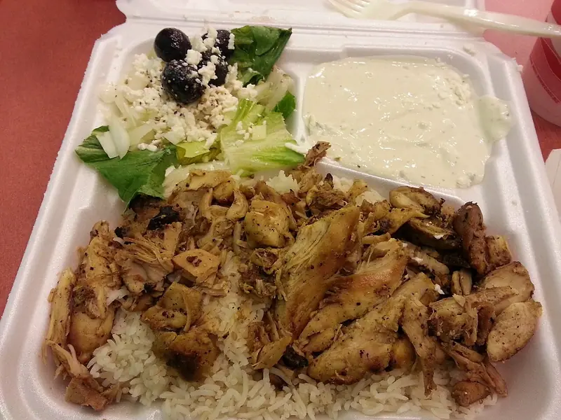 Takeout foam container of sliced seasoned chicken over white rice, a lettuce salad with feta and black olives, and white sauce