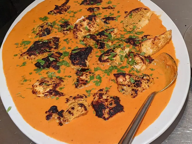 Charred chicken tikka pieces in a smooth orange curry sauce on a white platter, scattered with chopped coriander, with a serving spoon.