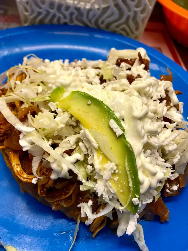 Kuřecí tinga na tostadě, posypaná nastrouhaným zelím, s cremou, rozdrobeným bílým sýrem a plátkem avokáda na modrém talíři