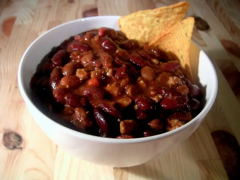 White bowl of chili with kidney beans and ground meat in a thick red sauce, with two tortilla chips tucked in, on a wooden table.