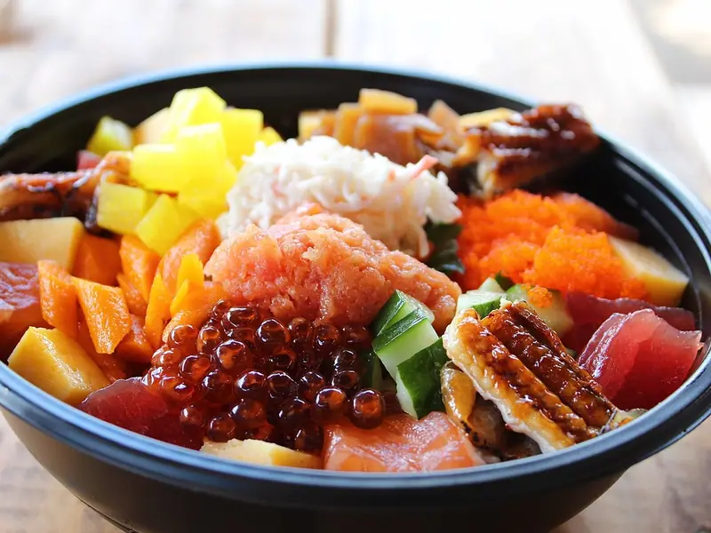 Chirashi-skål i en sort takeaway-boks toppet med tun, laks, lakserogn, tobiko, grillet ål, krabbesalat, tamago og agurk