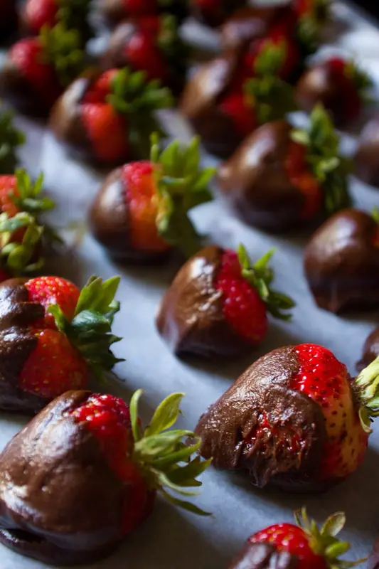 Rows of whole strawberries with green stems, partly dipped in dark chocolate, resting on parchment paper, with no visible toppings.