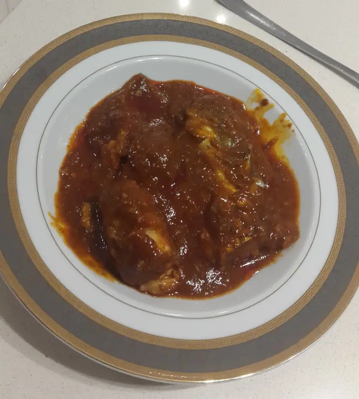 Stykker af hvid fisk dækket af en tyk, blank rødbrun tomatsauce, serveret i en hvid skål med grå- og guldmønstret kant.