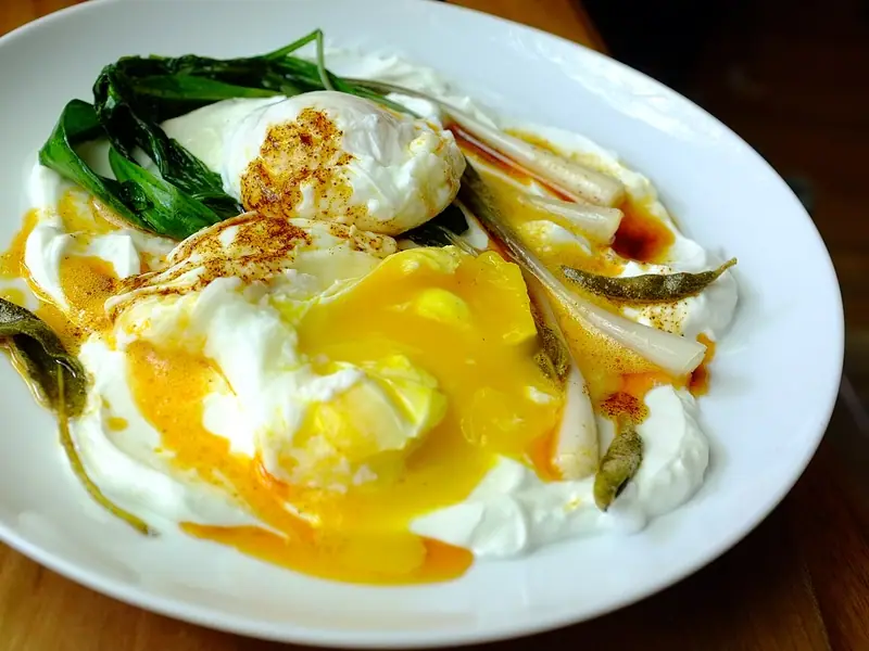 Two poached eggs, one cut open with runny yolk, on white yogurt with red pepper butter, wilted greens, spring onions and crisp sage leaves.