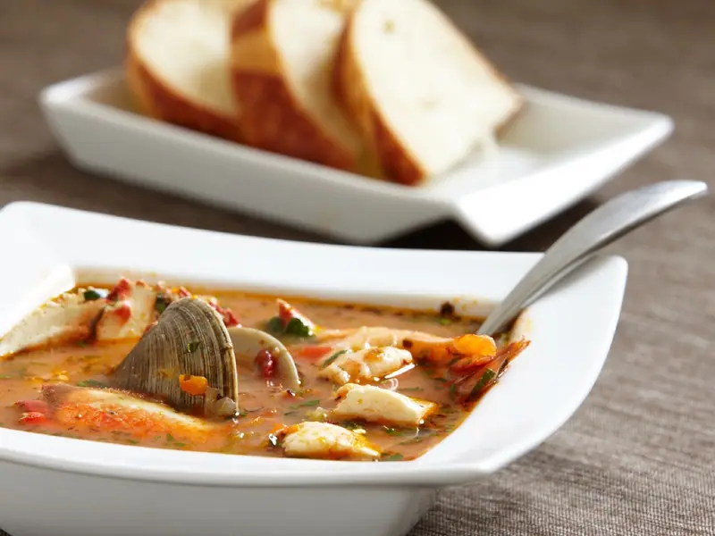 Tomato seafood stew with a clam, shrimp and white fish chunks in a square white bowl with a spoon, sliced bread on a plate behind