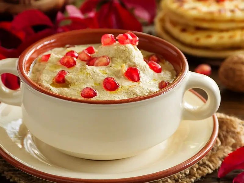 A cream ceramic bowl of smooth walnut dip topped with pomegranate seeds and olive oil, with flatbreads and red flowers behind.