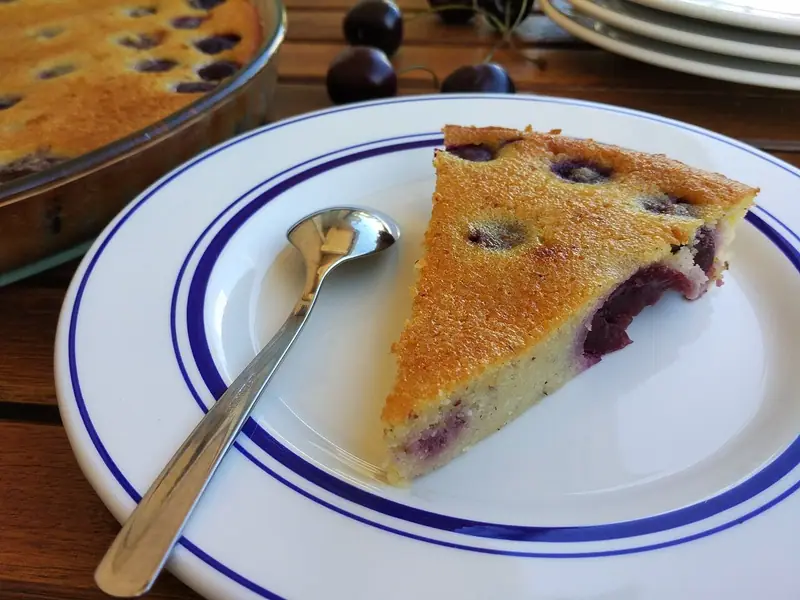 Et gyldent stykke kirsebærclafoutis på en hvid tallerken med blå kant og en ske, med bageformen og friske kirsebær i baggrunden.