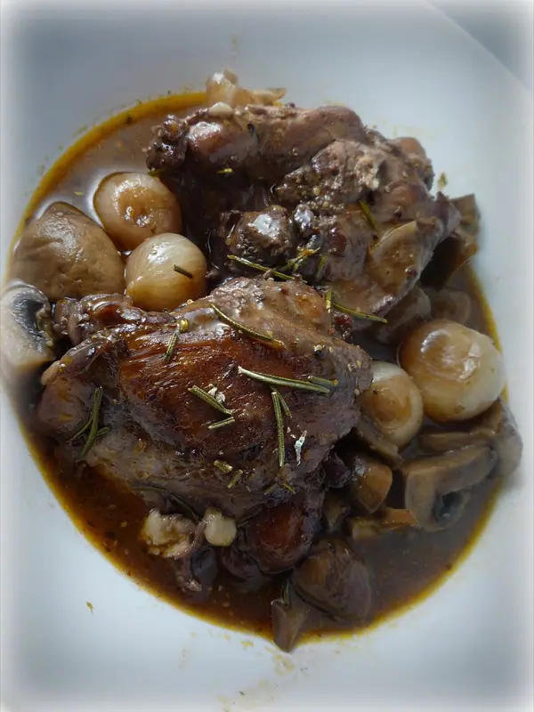 Braised dark meat pieces in a glossy brown wine sauce with pearl onions and mushrooms, sprinkled with rosemary, in a white bowl.