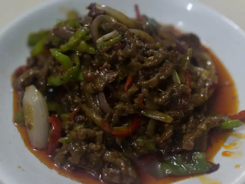 Shredded meat stir-fried with green chilli strips, red chilli, onion and a scallion piece in glossy red chilli oil on a white plate.