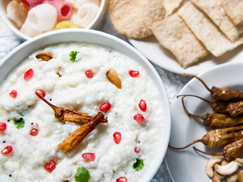 طبق أبيض من أرز باللبن الزبادي الكريمي مزيّن بحبوب الرمان وأوراق الكزبرة والفلفل المقلي، مع خبز الباباد والفلفل المقلي والكاجو