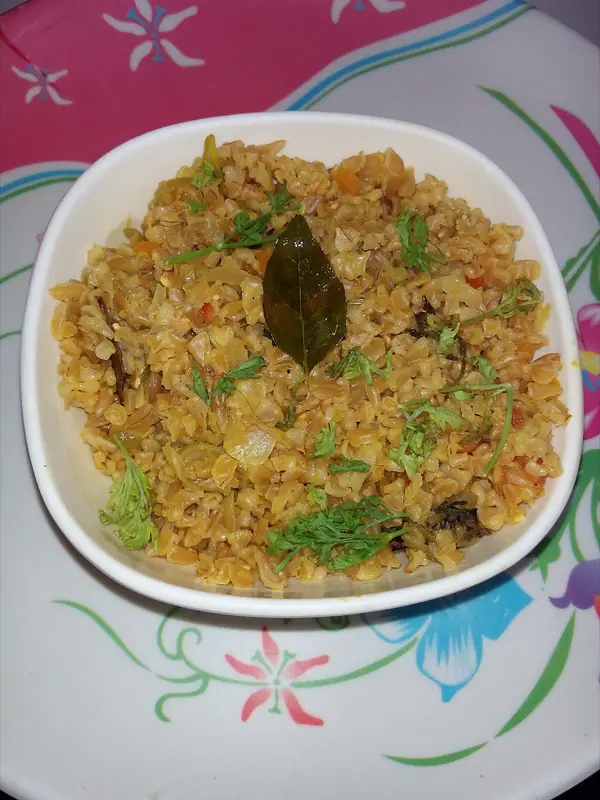 Fluffy spiced broken wheat with onion, cabbage and bits of tomato in a white square bowl, topped with coriander sprigs and a curry leaf.