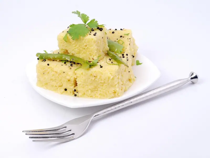 Gelbe Stücke gedämpfter Dhokla mit Senfsamen, grünen Chilis und Koriander auf einem weißen Teller neben einer Gabel.