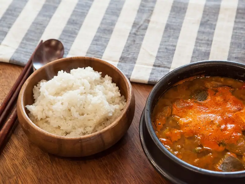Orangeroter koreanischer Eintopf mit Tofu und Gemüse in einem schwarzen Steintopf, daneben eine Holzschale mit gedämpftem Reis und Essstäbchen auf einem Holztisch