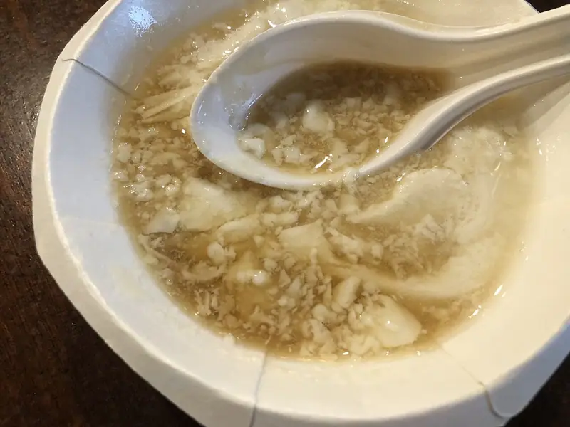 Beyaz kâğıt kâsede, açık altın renkli şurubun içinde ince dilimler ve ufalanmış parçalar hâlinde yumuşak beyaz tofu pudingi, yanında beyaz bir çorba kaşığı.
