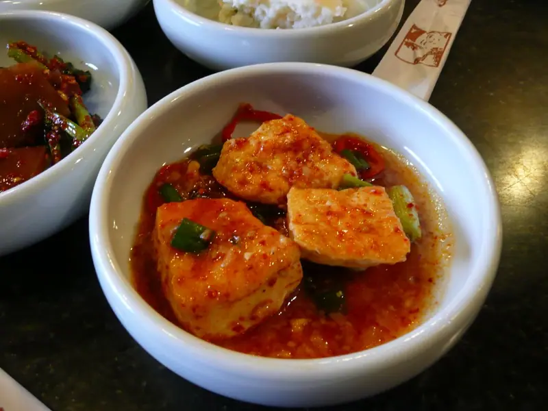 Tofubiter i rød chilisaus med vårløk og rød chili i en hvit bolle, med banchan-småretter og ris ved siden av.