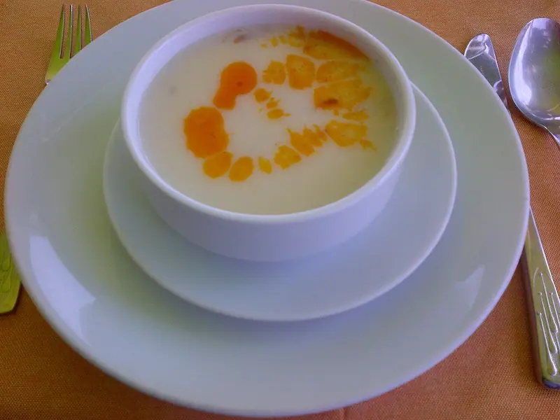 Cremige, helle Suppe in einer weißen Schale mit einem orangefarbenen Wirbel aus Paprikaöl, auf gestapelten weißen Tellern mit Gabel und Löffel auf einem orangefarbenen Tuch.