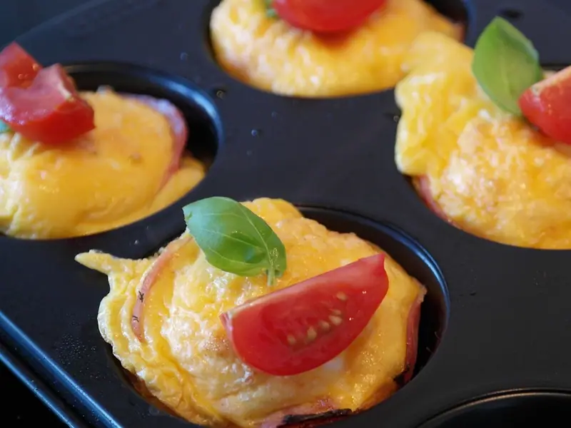 Gebackene Eiermuffins in einem schwarzen Muffinblech, auf Schinkenscheiben gesetzt und mit Kirschtomatenspalten und frischen Basilikumblättern belegt.
