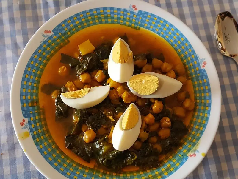 Guiso de garbanzos y espinacas en salsa anaranjada, coronado con cuatro cuñas de huevo duro, en un cuenco con borde azul sobre un mantel de cuadros.