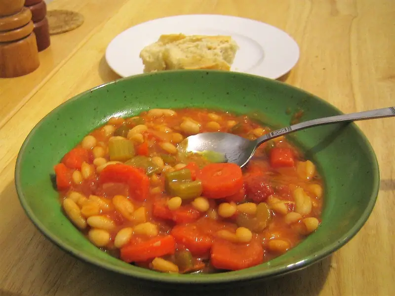 Tomato white bean soup with carrot rounds and celery in a green bowl with a spoon, crusty bread on a white plate behind