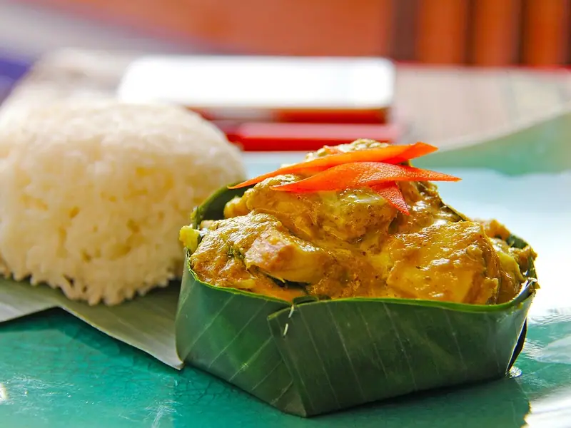 Fish amok in yellow coconut curry served in a folded banana-leaf cup, topped with red chilli strips, beside a mound of white rice.