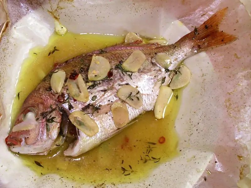 A whole roasted red-skinned fish topped with sliced garlic, thyme and chili flakes, sitting in golden olive oil on opened parchment.