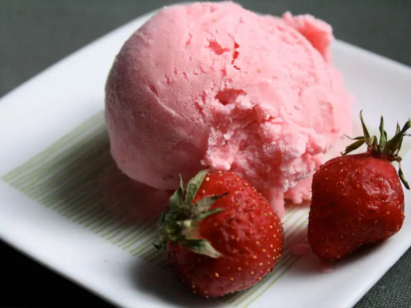 A scoop of pink strawberry frozen dessert on a white square plate with green stripes, beside two whole fresh strawberries.