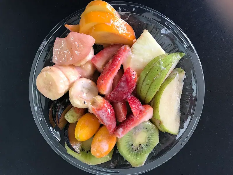 Clear plastic bowl of cut fruit including strawberries, banana, kiwi, pear, plum, kumquats, grapefruit and orange on a dark surface.