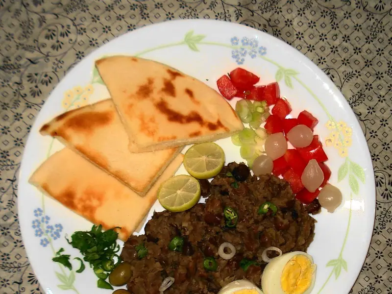 Haşlanmış yumurta yarımları, yeşil zeytin, yarım misket limonları, küp doğranmış domates, soğan turşusu, taze otlar ve pide dilimleriyle servis edilmiş bir tabak ezilmiş bakla.