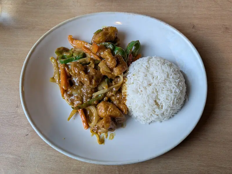 Kurczak w panierce w złocistym sosie w stylu curry z paskami zielonej papryki, marchewki i cebuli obok kopczyka białego ryżu na białym talerzu.