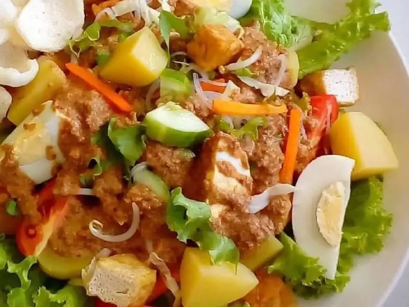 Gado-Gado auf Salatblättern in einer weißen Schüssel mit Kartoffeln, Tofu, gekochtem Ei, Gurke, Karotte und Erdnusssoße, dazu Krabbenchips.