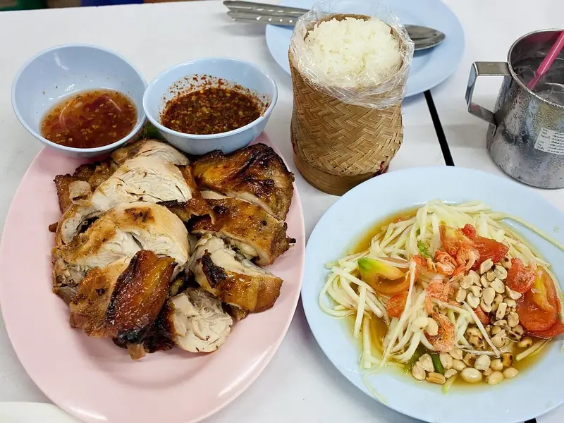 Grilled chicken pieces on a pink plate with two chilli dipping sauces, green papaya salad with peanuts, and sticky rice in a basket