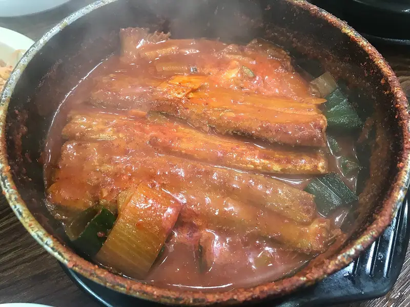 Steaming cutlassfish pieces braised in red chili sauce with onion and green onion in a black stone pot, crusted with chili at the rim.