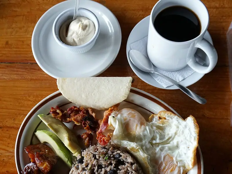 Gallo pinto med to spejlæg, avocadoskiver, bacon, stegte plantains og en tortilla, med creme fraiche og sort kaffe ved siden af