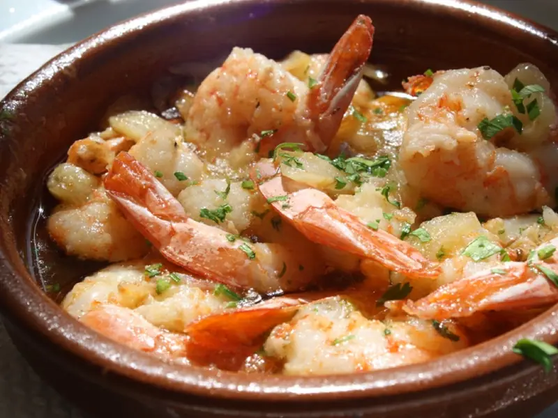 Shrimp with tails on in golden garlic oil, scattered with chopped parsley, served in a brown clay cazuela dish.