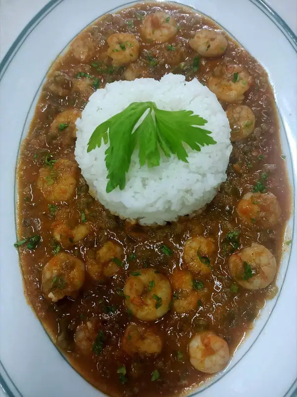 Rejer i en tyk rødbrun sauce med hakket persille på et ovalt hvidt fad, ved siden af en bunke hvide ris toppet med et selleriblad.