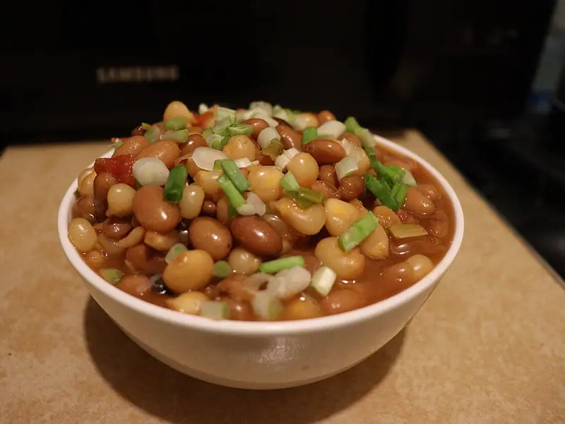 White bowl of pinto-style beans and yellow maize kernels in a light tomato sauce, topped with chopped green onions, on a tan board.