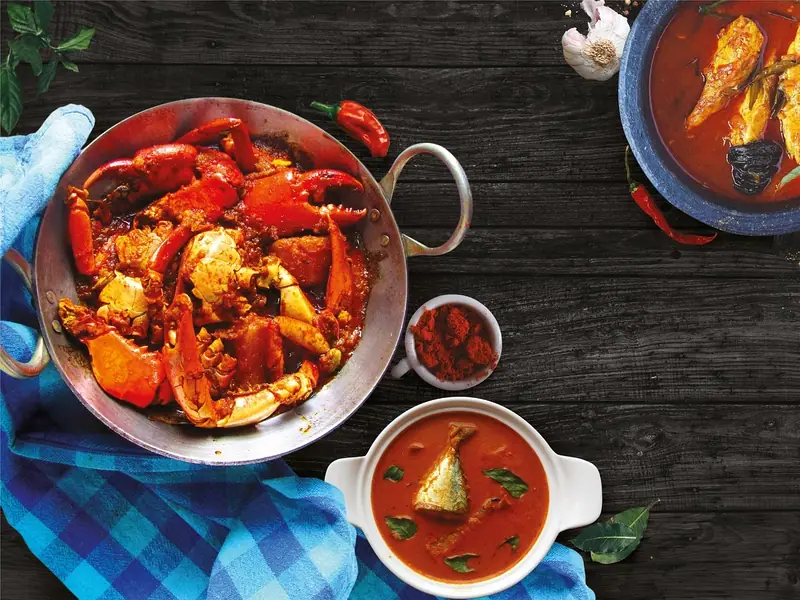 Červené krabí kari v kovové pánvi vedle misek s červeným rybím kari, kari listy a chilli práškem na tmavém dřevěném stole.