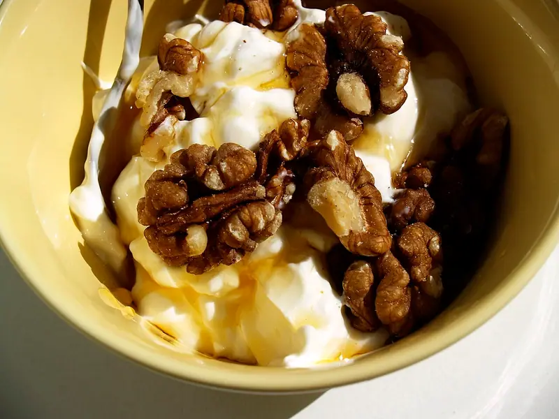 Yellow bowl of thick white yogurt topped with whole walnut halves and a drizzle of golden honey, in bright sunlight.
